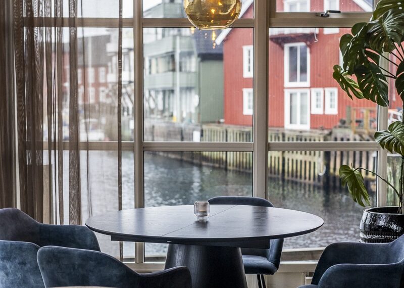 Rundt bord og blå stoler ved vindu mot kanal i Henningsvær